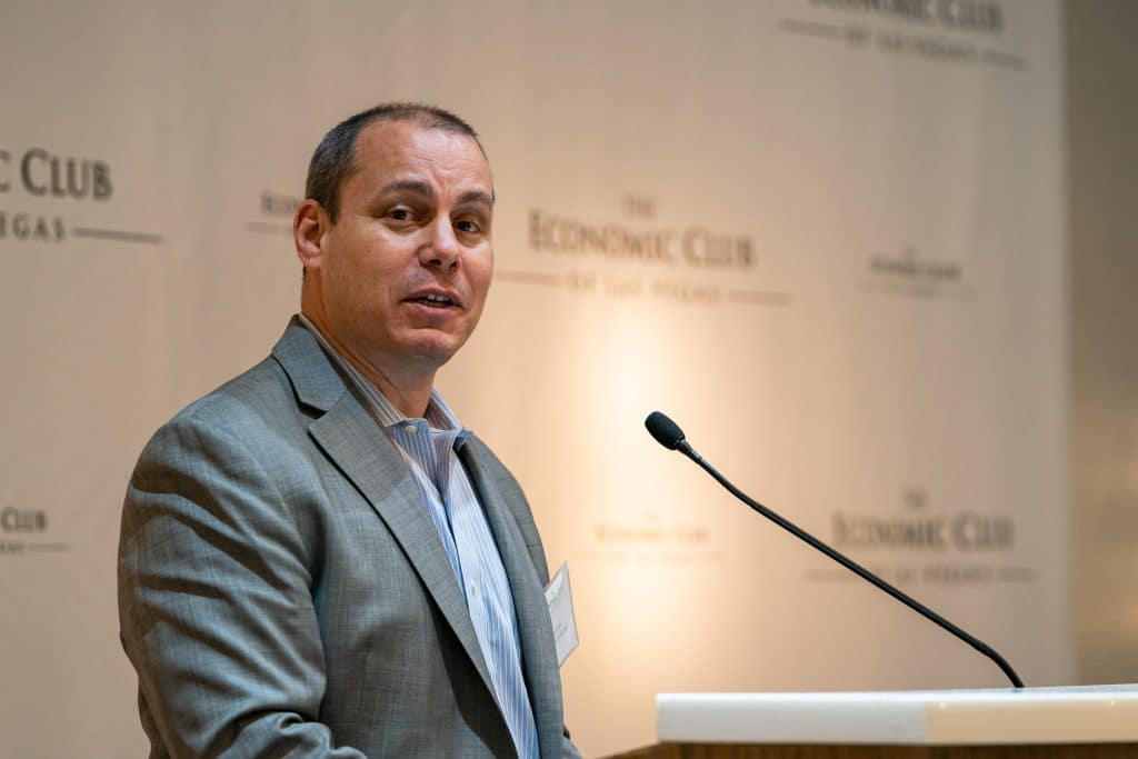 A man in a gray suit and blue striped shirt speaks at a podium with "THE ECONOMIC CLUB" on the wall.