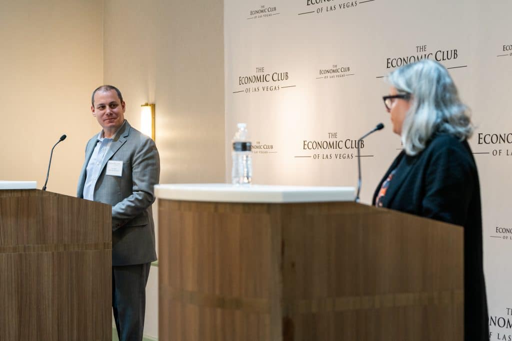 A man in a suit stands at a podium, smiling, while a woman with gray hair speaks at another podium.