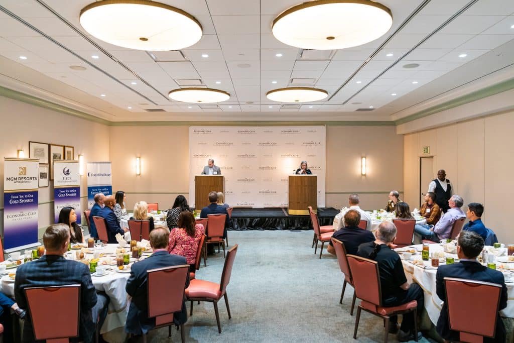 A large conference room has an audience seated at round tables facing two speakers at podiums.