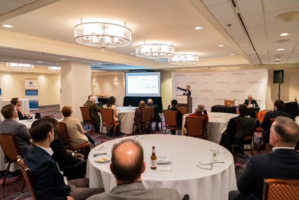 A speaker at a podium gestures toward a screen showing a graph, while an audience sits at round tables.