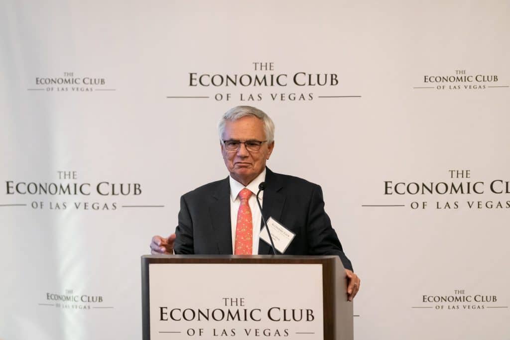 A man in a suit and red tie speaks at a podium with "The Economic Club of Las Vegas" logo.