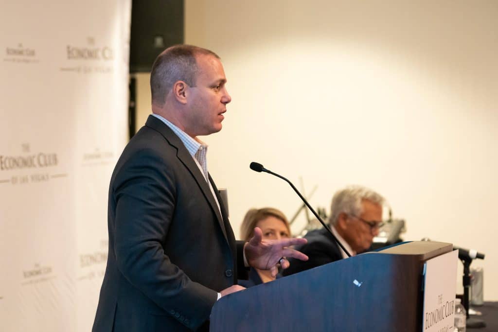 A man in a suit speaks at a podium with a microphone, gesturing with his right hand. "The Economic Club" is visible on the backdrop.