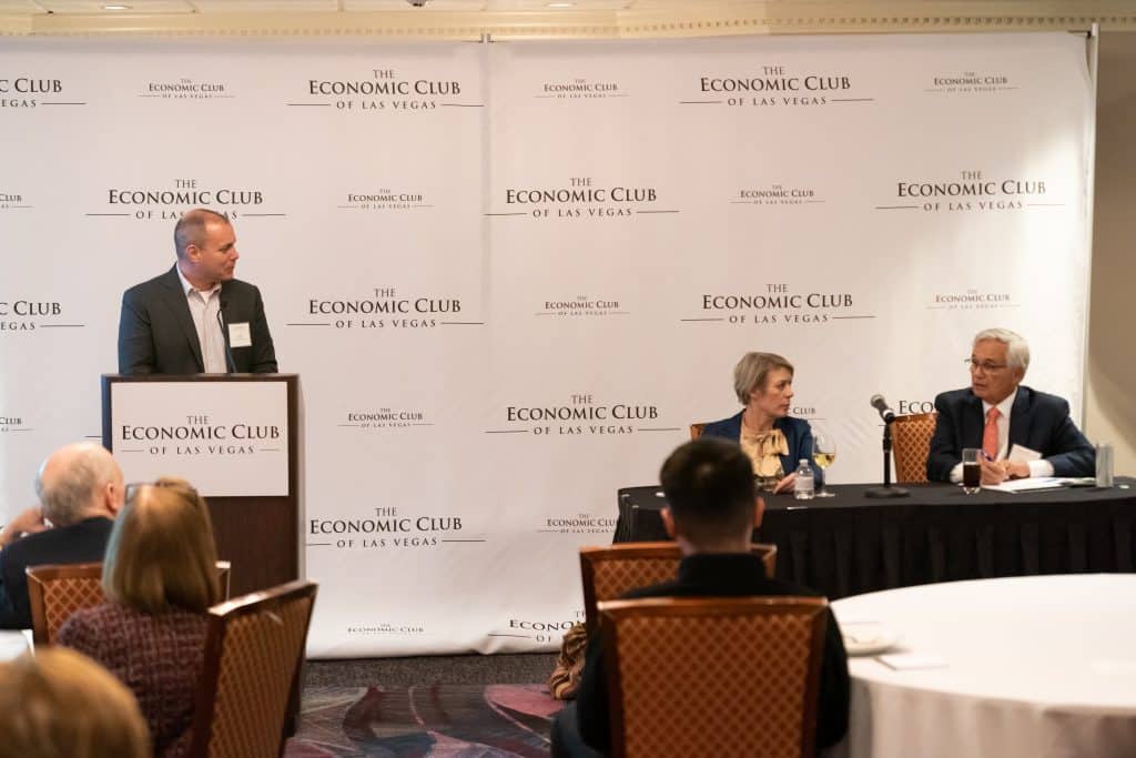 A man speaks at a podium while a woman and man sit at a table in front of a "The Economic Club of Las Vegas" backdrop.