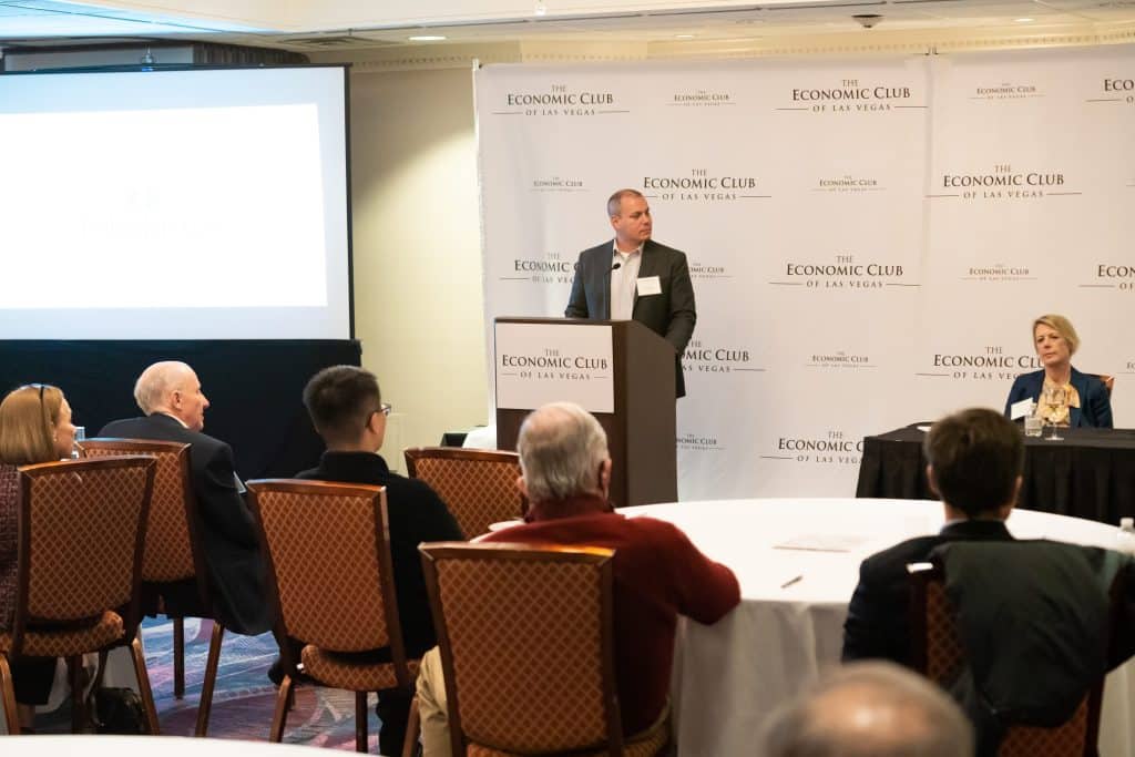 A man speaks at a podium with "The Economic Club of Las Vegas" repeated on the backdrop, while attendees listen.