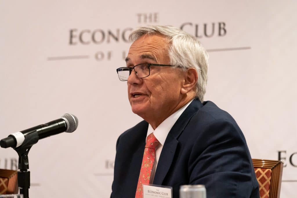 A gray-haired man in glasses and a dark suit speaks into a microphone, with "THE ECONOMIC CLUB" visible behind him.