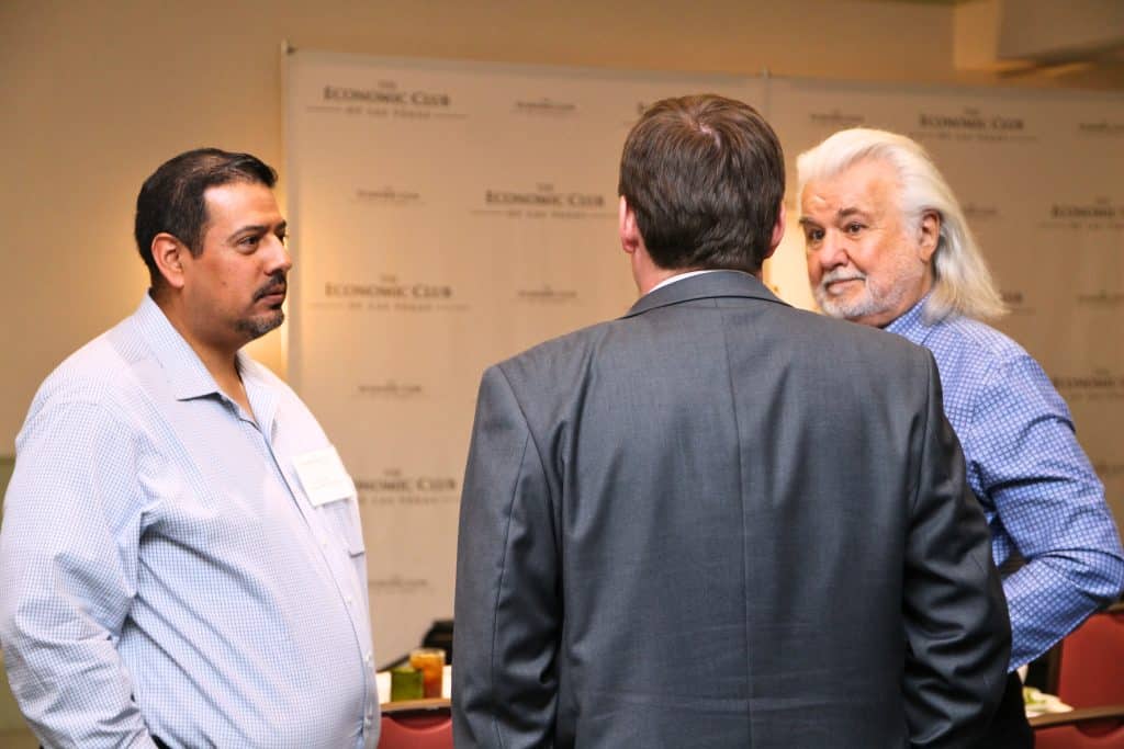 Three men stand in a room with "THE ECONOMIC CLUB" repeated on the wall behind them.