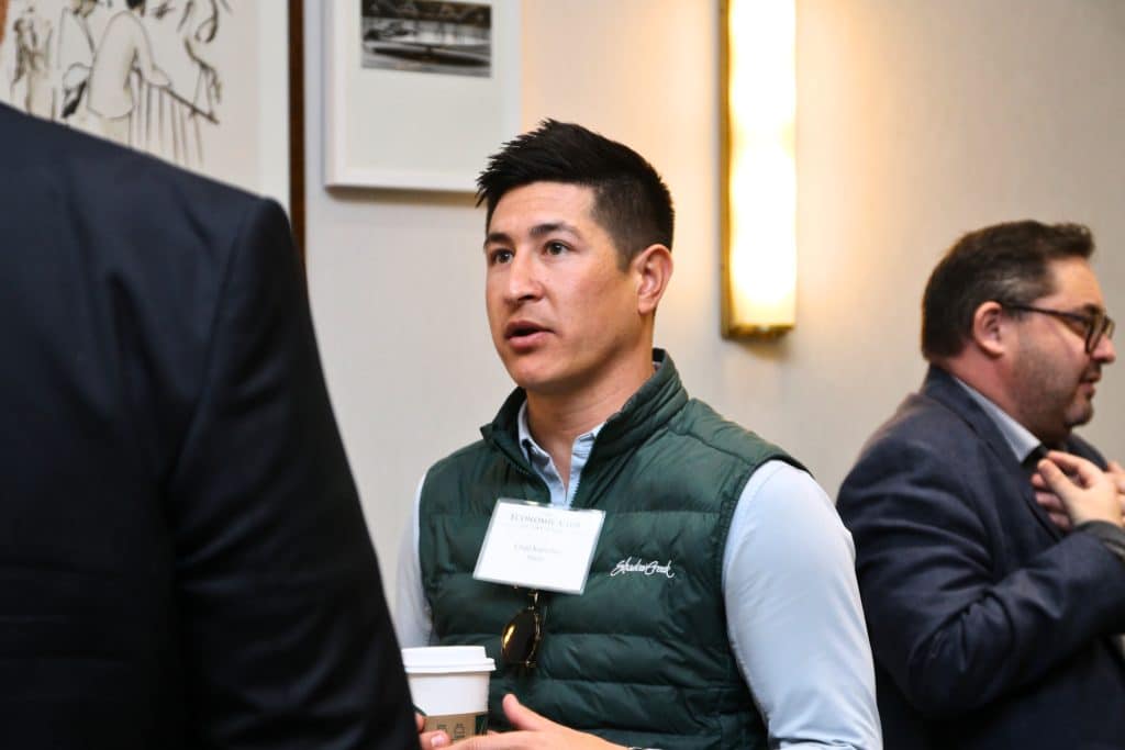 A man in a green vest and light blue shirt speaks, holding a coffee cup and wearing a name tag.