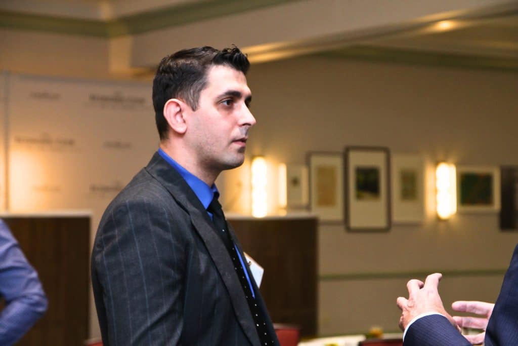 A man with dark hair and a blue shirt, wearing a pinstripe suit, looks right in a room with framed art.