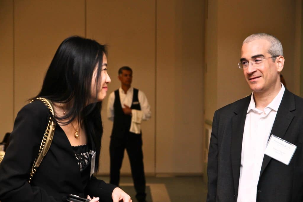A woman with long dark hair smiles at a man in glasses and a suit, with a waiter in the background.