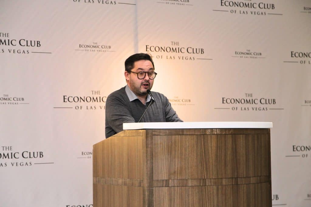 A man with glasses and a gray sweater speaks at a wooden podium in front of a wall with "THE ECONOMIC CLUB OF LAS VEGAS" repeated.