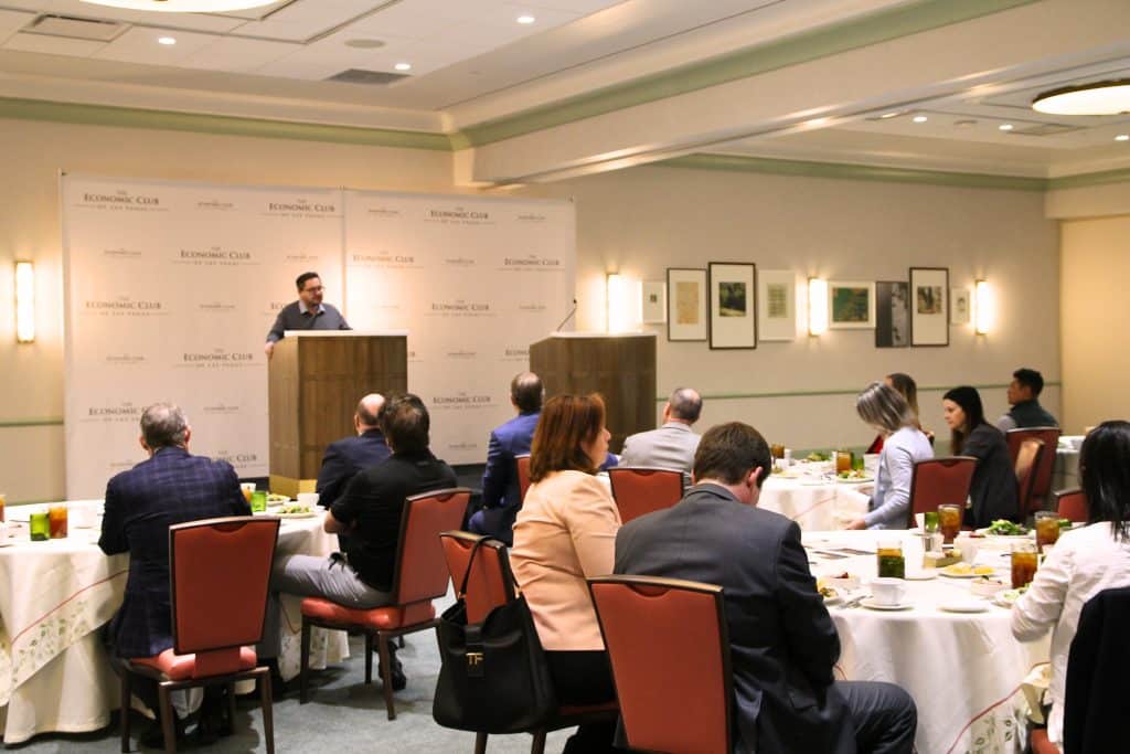 A man speaks at a podium in front of a "The Economic Club" backdrop to an audience seated at round tables.