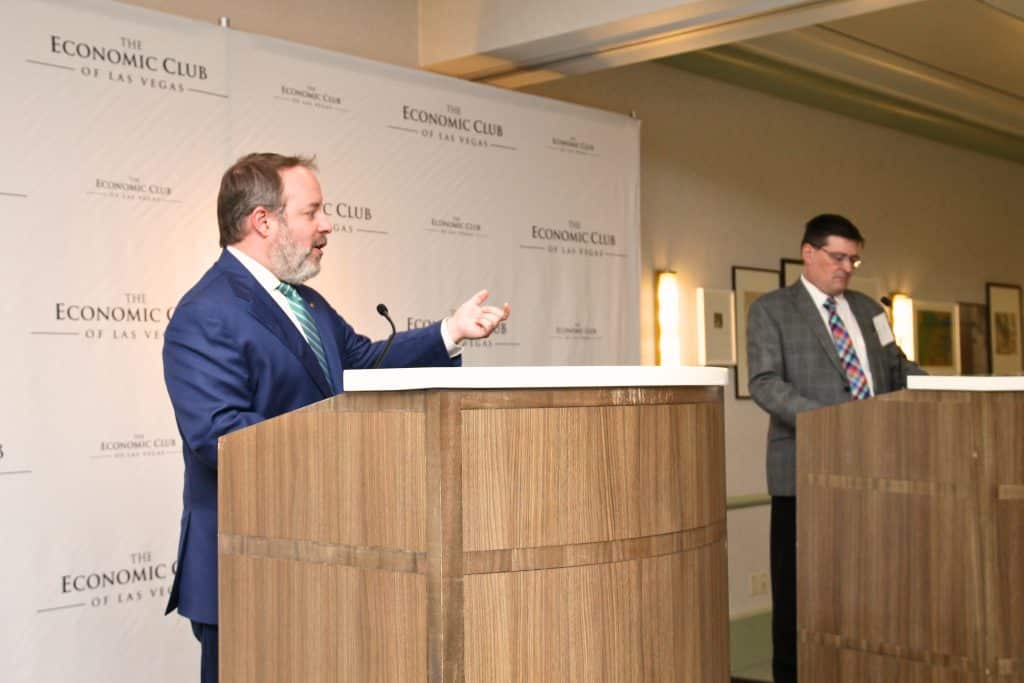 A bearded man in a blue suit speaks from a podium, gesturing, with "THE ECONOMIC CLUB OF LAS VEGAS" banners behind him.