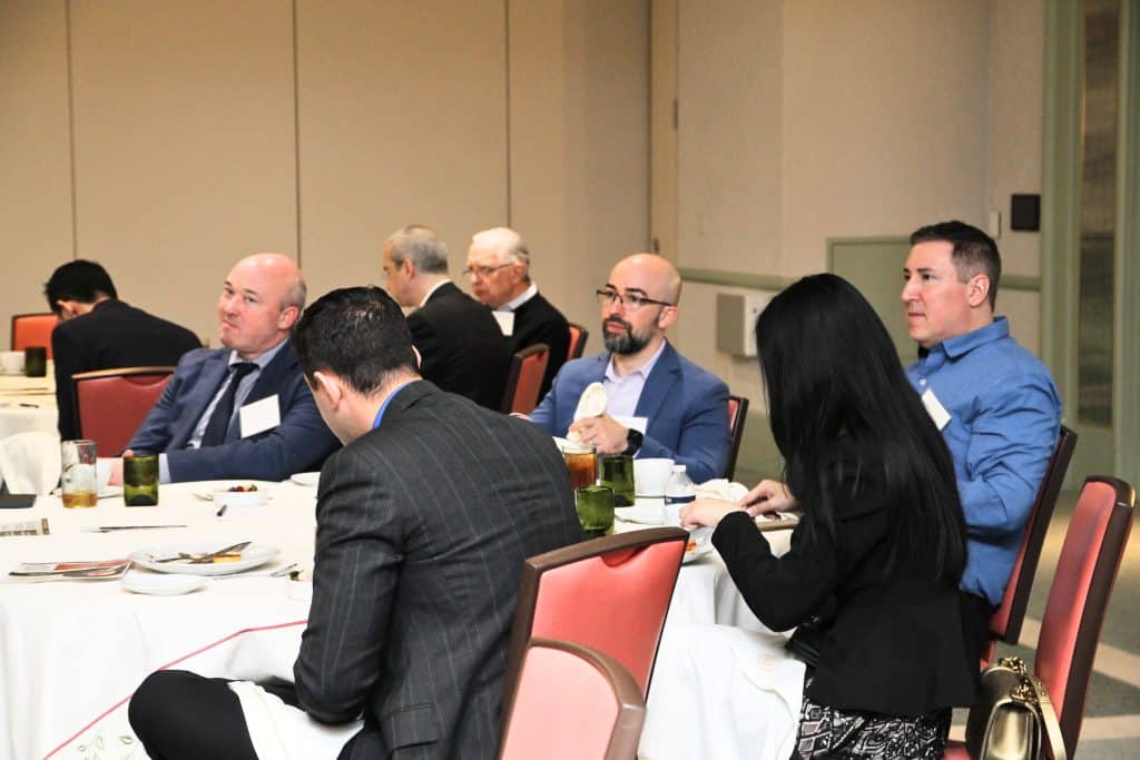 A diverse group of professionals sits at round tables with white tablecloths, listening intently during a conference.