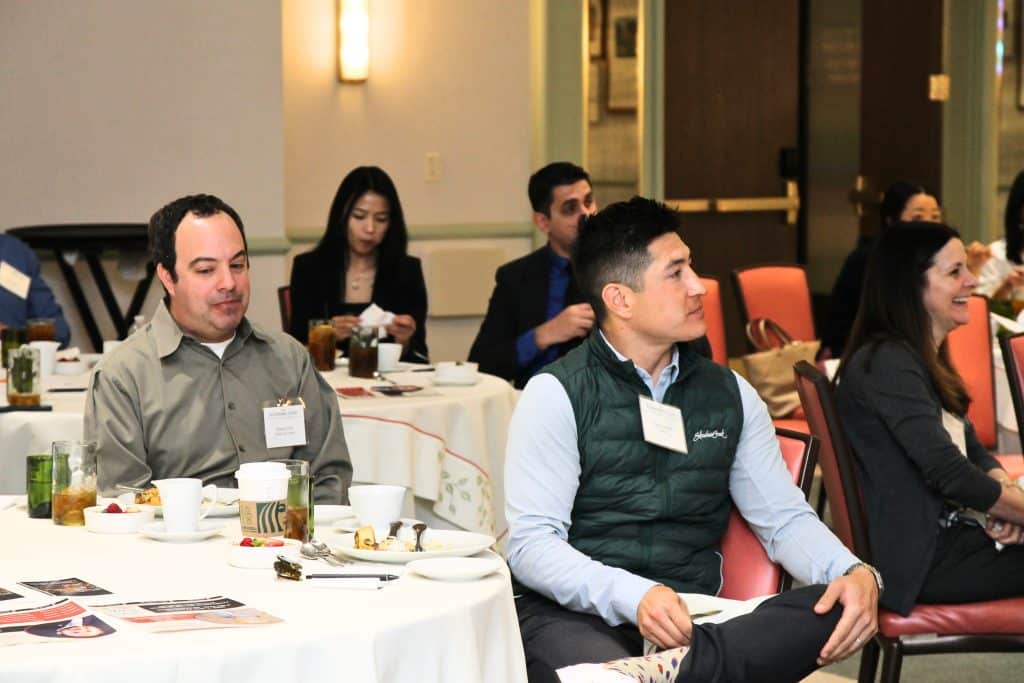 Attendees sit at round tables with drinks and food, listening to a presentation in a brightly lit room.