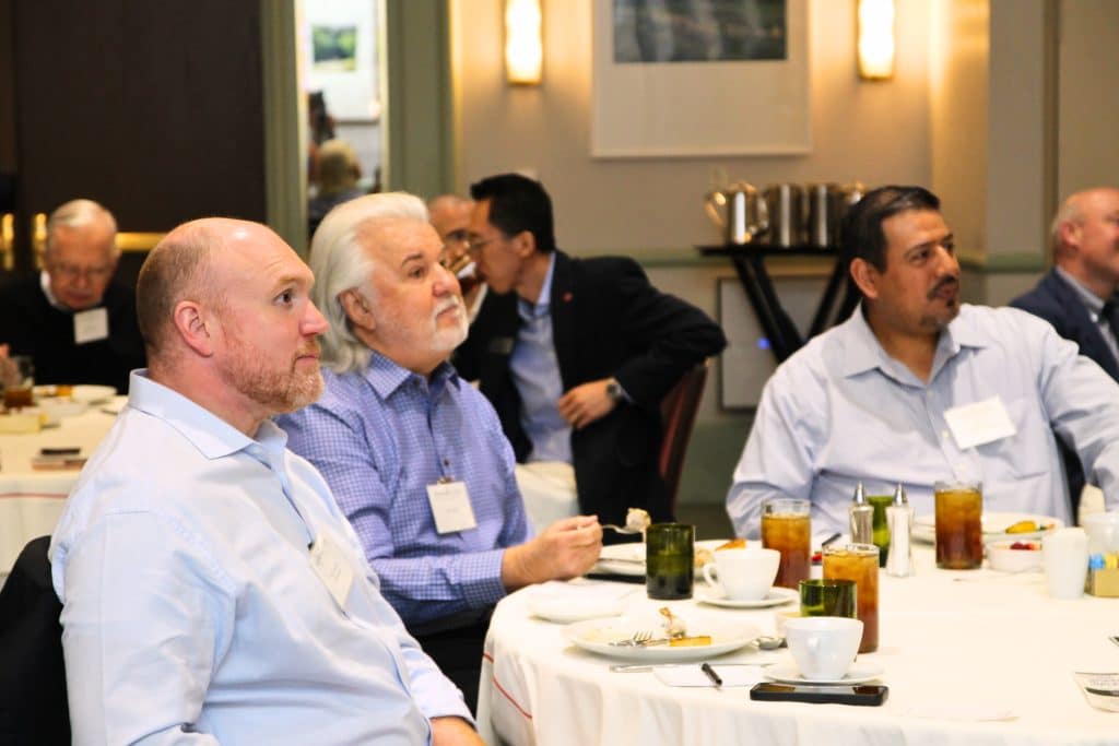 Men with name tags are seated at a white-clothed table with drinks and plates in a conference room.
