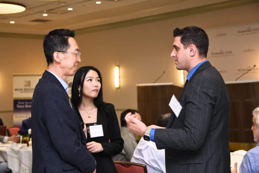 Three people, two men and one woman, wearing business attire and name tags, converse at a conference.