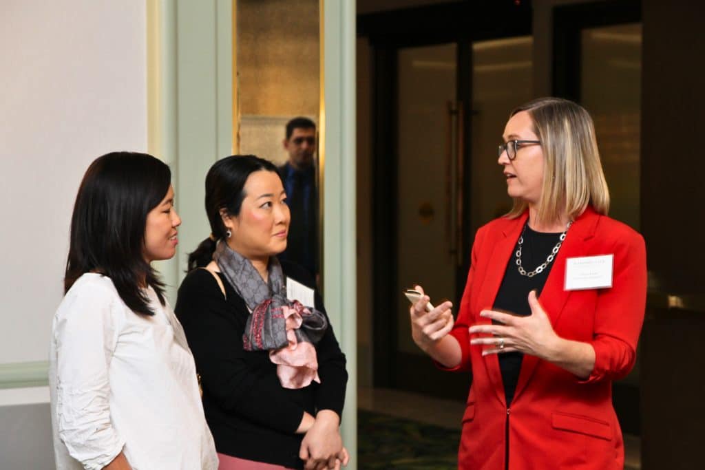 Three women are conversing; one in a red blazer gestures while two others listen intently.