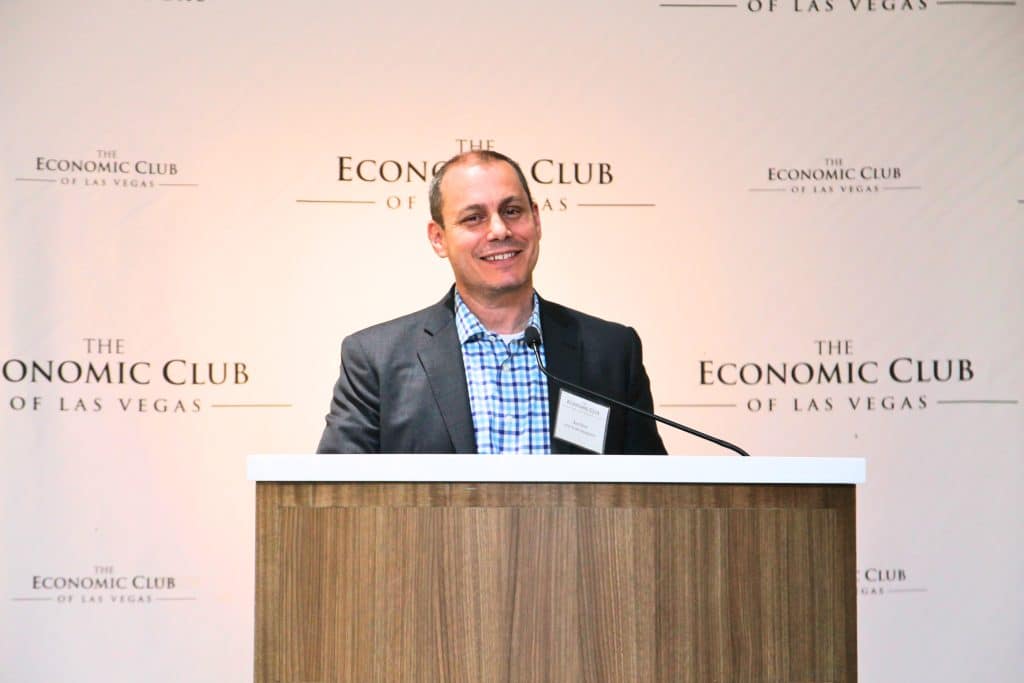 A smiling man in a suit and plaid shirt stands behind a podium with "The Economic Club of Las Vegas" on the wall.
