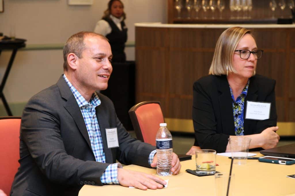 A man in a blue plaid shirt and suit jacket smiles next to a woman in glasses and a black blazer.