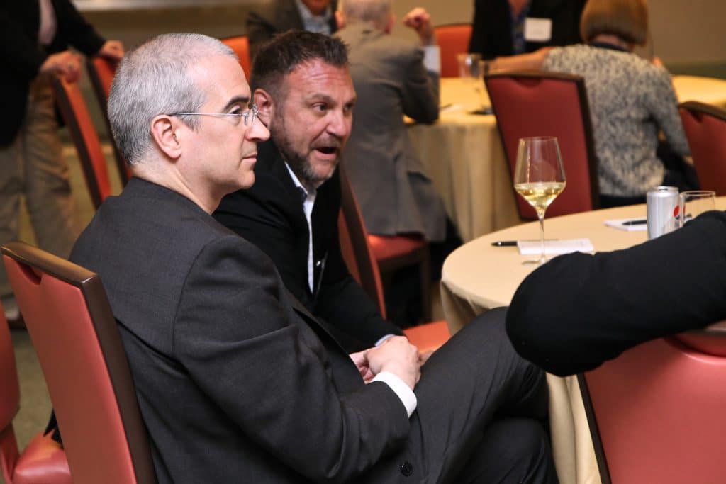 Two men in suits sit at a table with drinks; one has gray hair and glasses, the other a beard.
