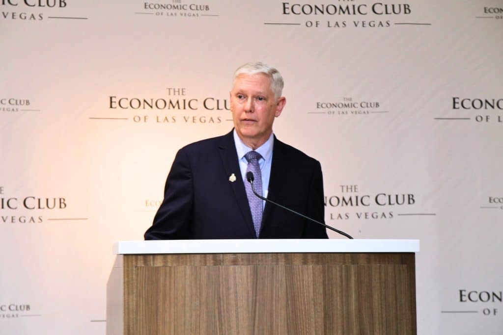 A man in a suit and tie speaks at a wooden podium with "The Economic Club of Las Vegas" on the wall.