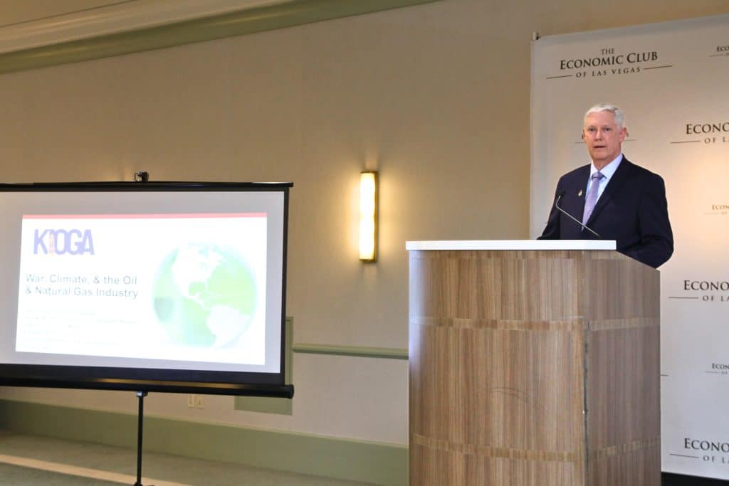 A man in a suit speaks at a podium, next to a screen displaying "KIOGA War, Climate, & The Oil & Natural Gas Industry."