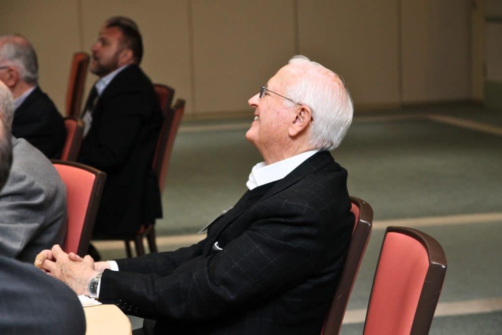A smiling, white-haired man in a suit and glasses sits in a red chair, looking up and to the left.