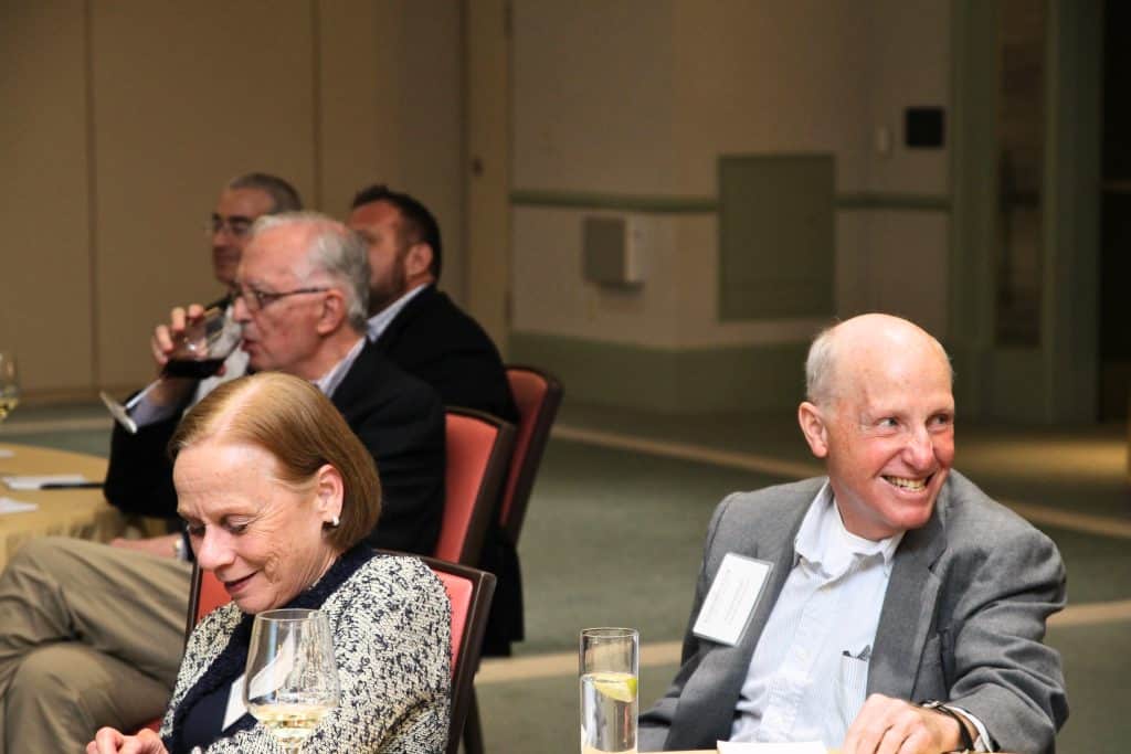 A group of people sitting at tables, with a man in a gray suit smiling and a woman looking down.