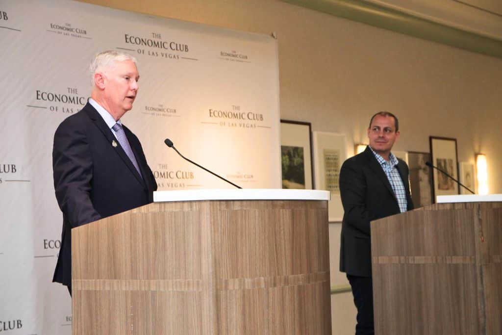 A man in a suit speaks at a podium with "The Economic Club of Las Vegas" on the backdrop, another man stands right.