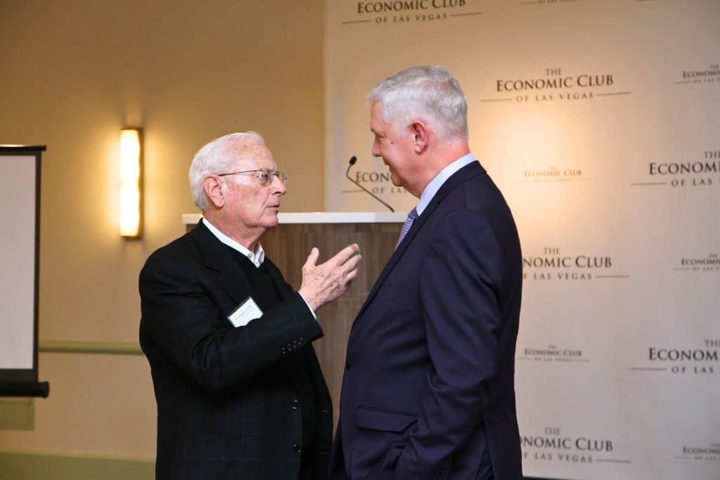 Two men in suits stand talking, with "THE ECONOMIC CLUB OF LAS VEGAS" repeated on the wall behind them.