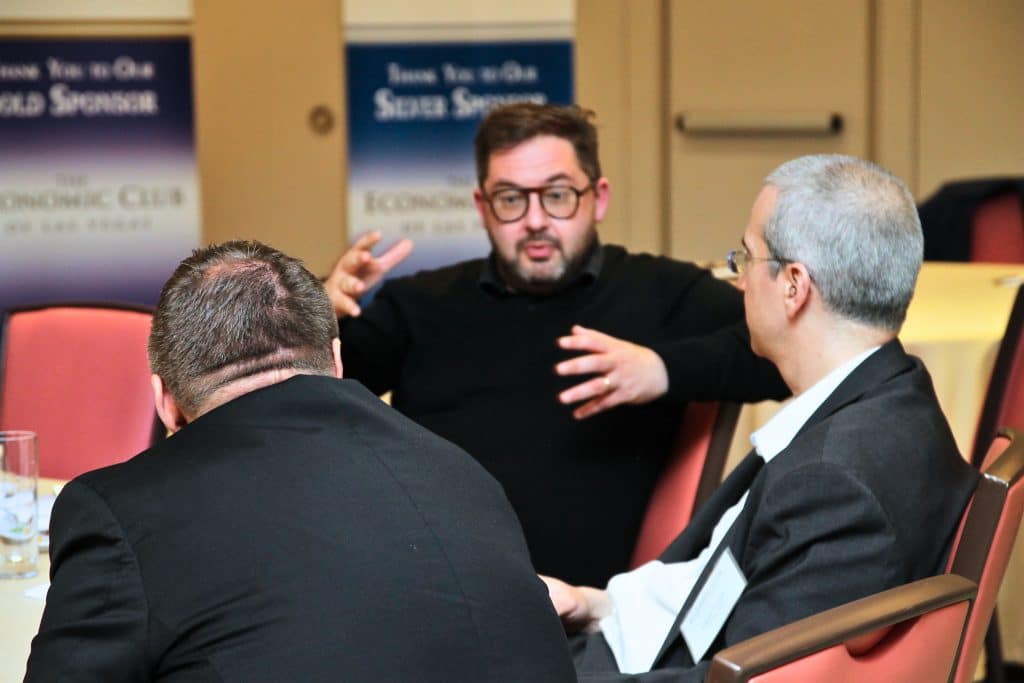 A bearded man in glasses and a black sweater gestures while talking to two men with their backs to the camera.