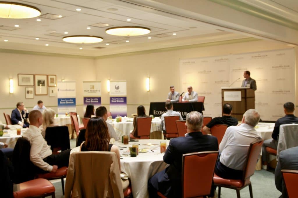 A speaker at a podium addresses an audience seated at round tables in a conference room with "ECONOMY CLUB" banners.
