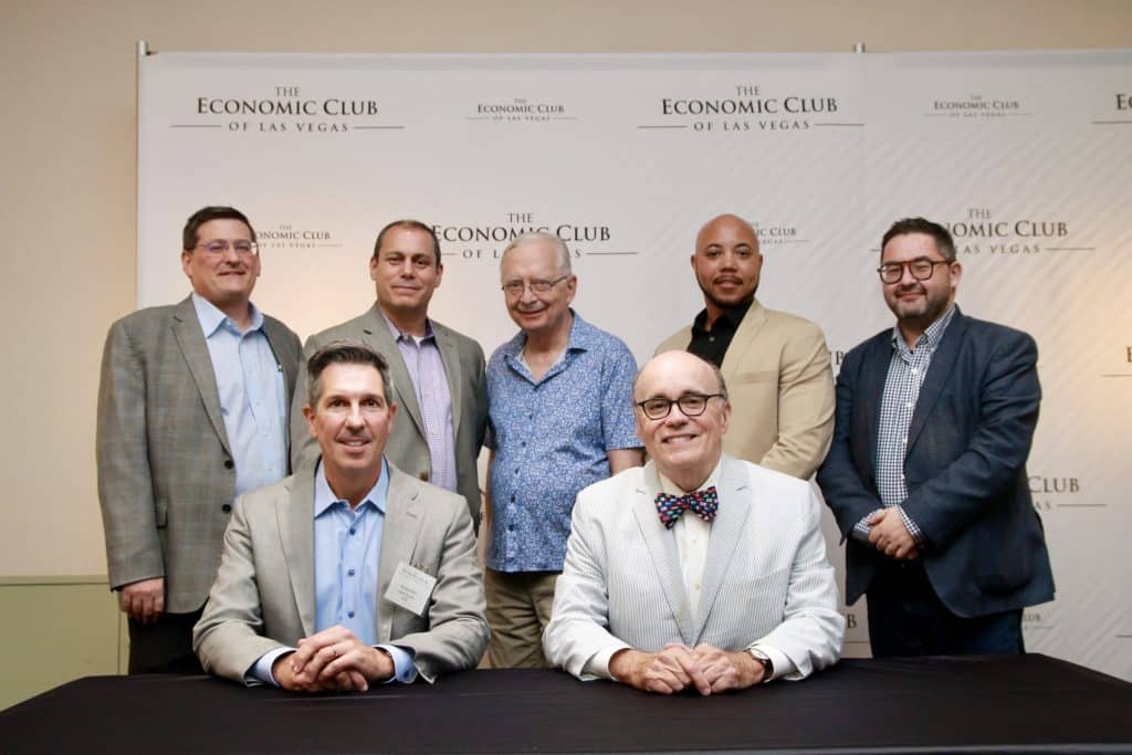 Seven men smile at the camera in front of a white wall with "The Economic Club of Las Vegas" repeated.