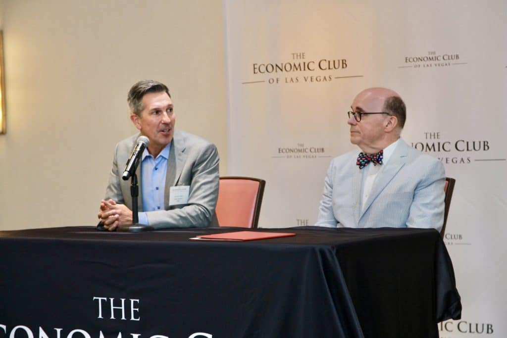 Two men sit at a black-draped table in front of a wall with "The Economic Club of Las Vegas" repeated.
