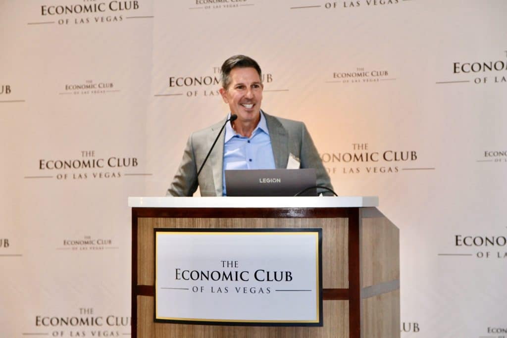 A smiling man in a gray suit and blue shirt speaks at a podium with "THE ECONOMIC CLUB OF LAS VEGAS" logo.