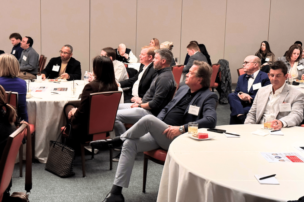 Attendees are seated at round tables with white tablecloths, listening intently to a presentation.