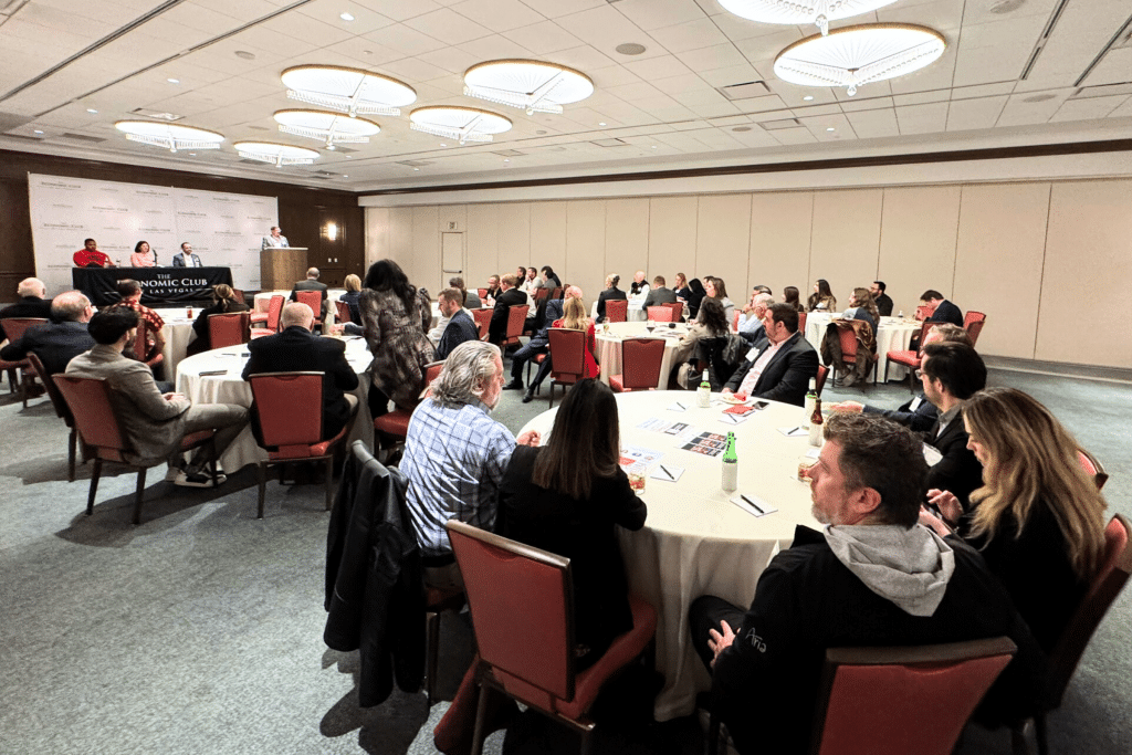 A large conference room with many people seated at round tables, facing a panel on a stage with "THE ECONOMIC CLUB LAS VEGAS" banner.