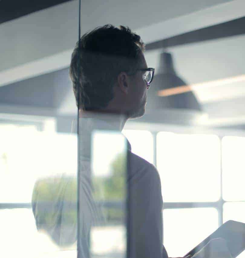 A man with dark hair and glasses looks right, partially obscured by a glass panel reflecting a window.